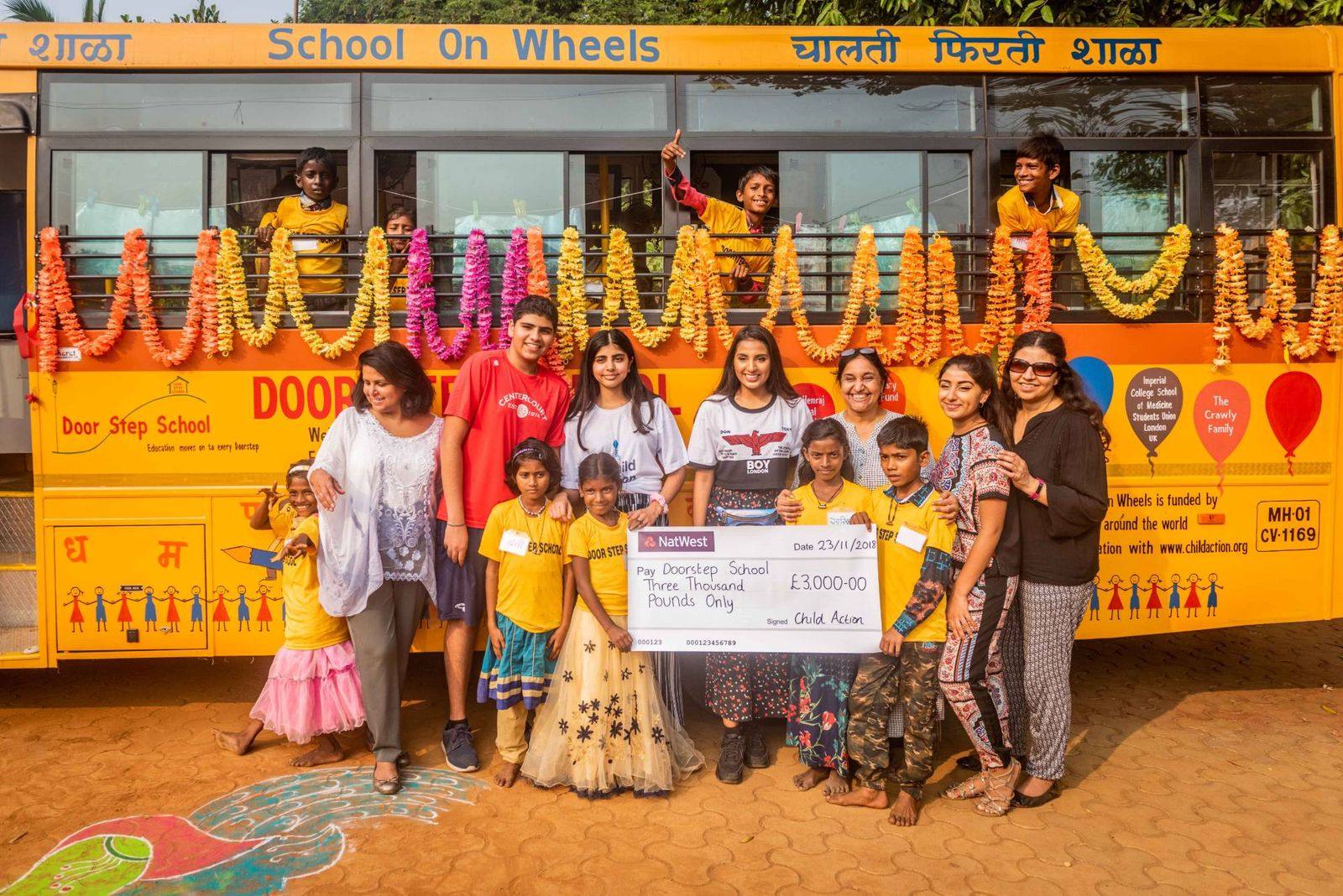 The youth ambassadors, Seema and Doorstep School children presenting a £3,000 cheque in front of a garlanded School on Wheels bus