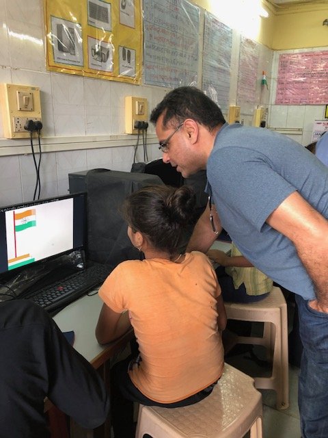 A visitor leaning over to watch a girl working at a computer in the Door Step School computer centre