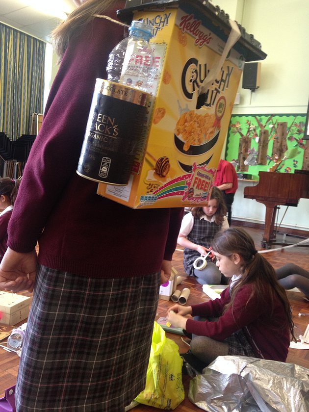 A pupil wearing a backpack made from a cereal box and plastic bottles while classmates build in the background